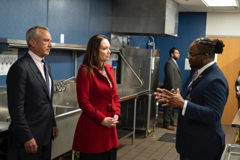 A school staff person talking to Secretary Rollins and Secretary Kennedy in the school cafeteria kitchen.