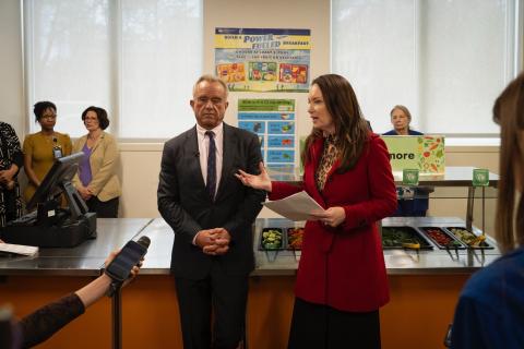 Secretary Rollins answering reporter questions in front of a school cafeteria salad bar alongside Secretary Kennedy.