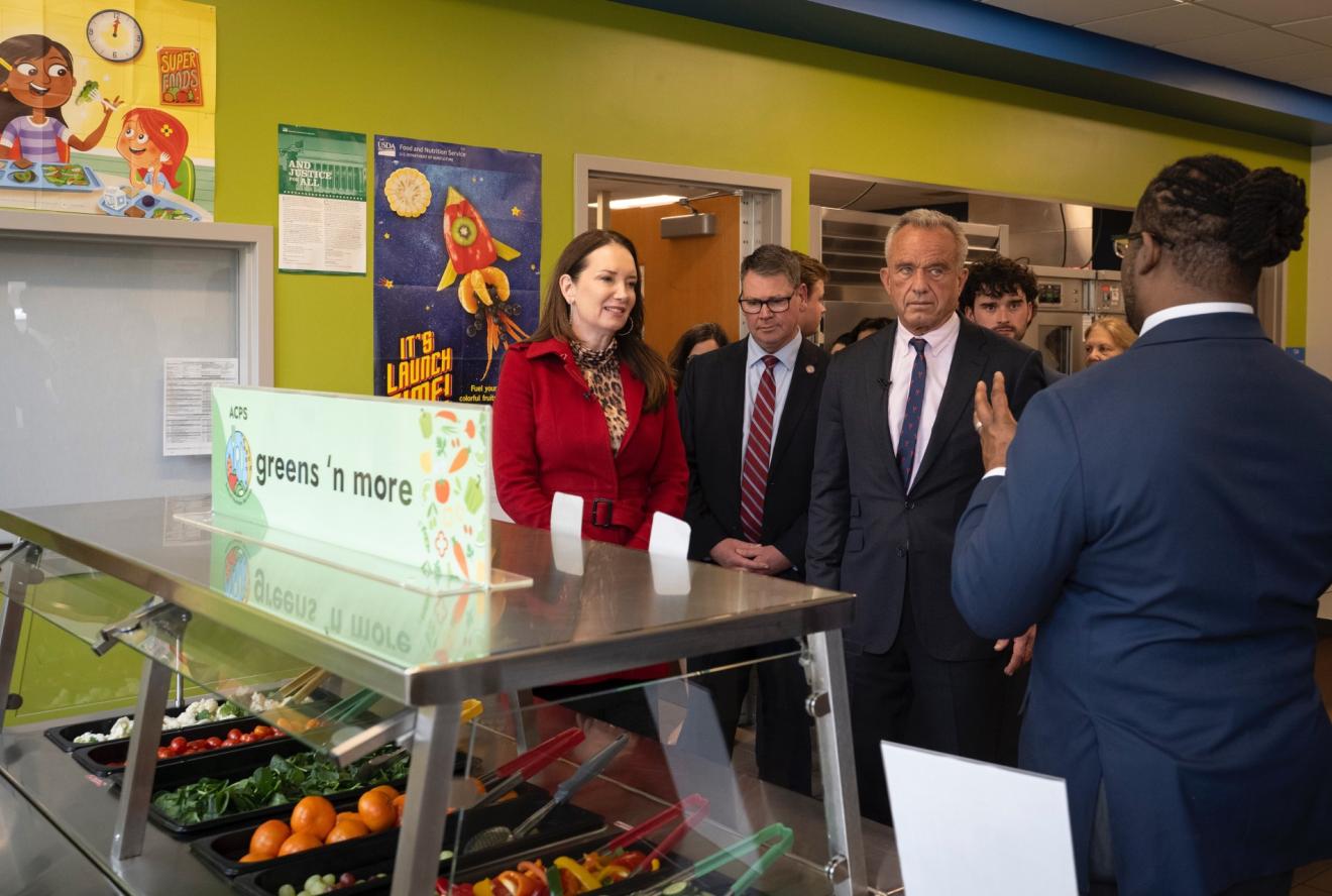 USDA and HHS secretaries at MAHA event in front of a salad bar in a school cafeteria.