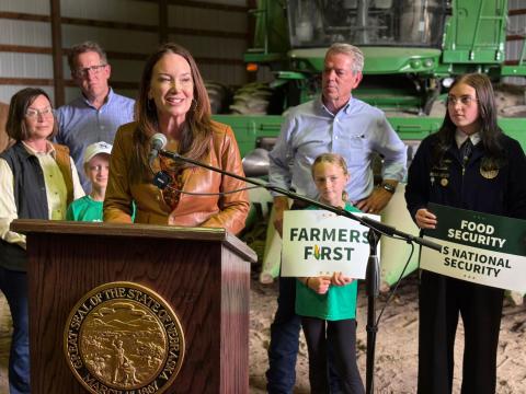Secretary Rollins at a podium speaking with a family behind her holding signs saying Farmers First.