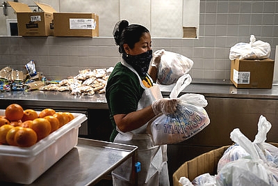 a school nutrition professional wearing a mask carrying bagged lunches next to a bin of oranges