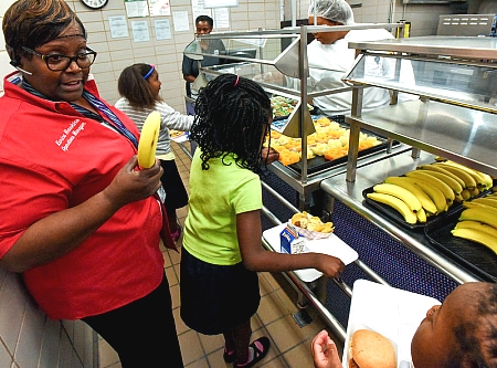 a school nutrition professional in a red uniform holding a banana and speaking to kids moving through a school cafeteria lunch line