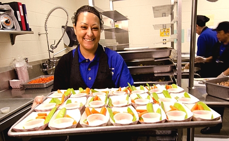a school nutrition professional wearing a hairnet and holding a tray of vegetables smiling