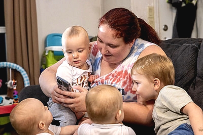 a woman looking at her phone holding an infant and surrounded by 3 toddlers