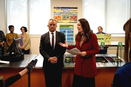 Secretary Rollins answering reporter questions in front of a school cafeteria salad bar alongside Secretary Kennedy.